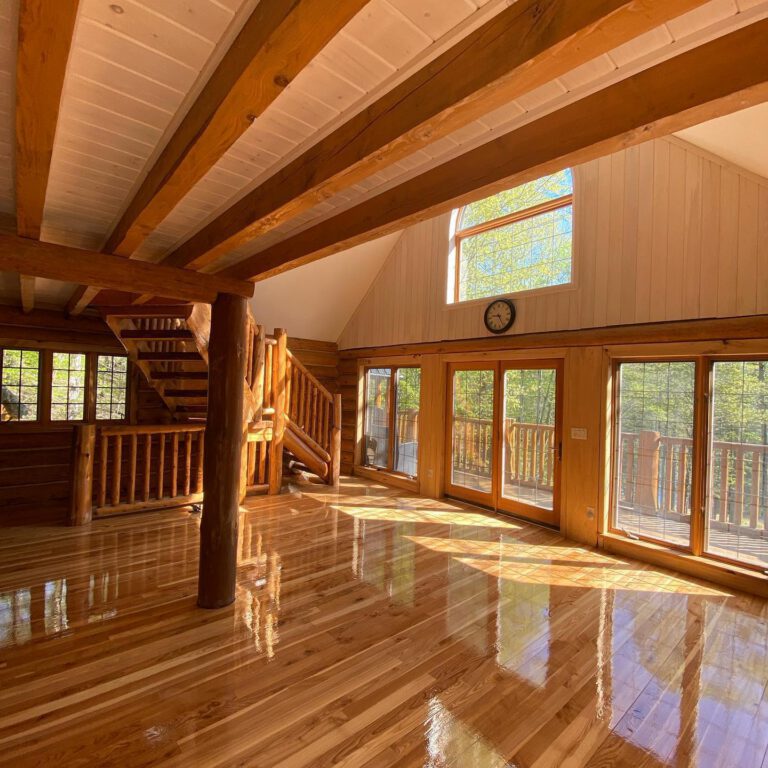 Freshly refinished hardwood floor in an open-plan log home interior, with exposed ceiling beams, a log staircase and patio doors leading onto a deck