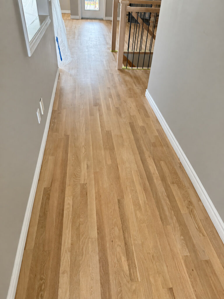 Light oak strip flooring running down an upstairs hallway past a stair railing with black balusters