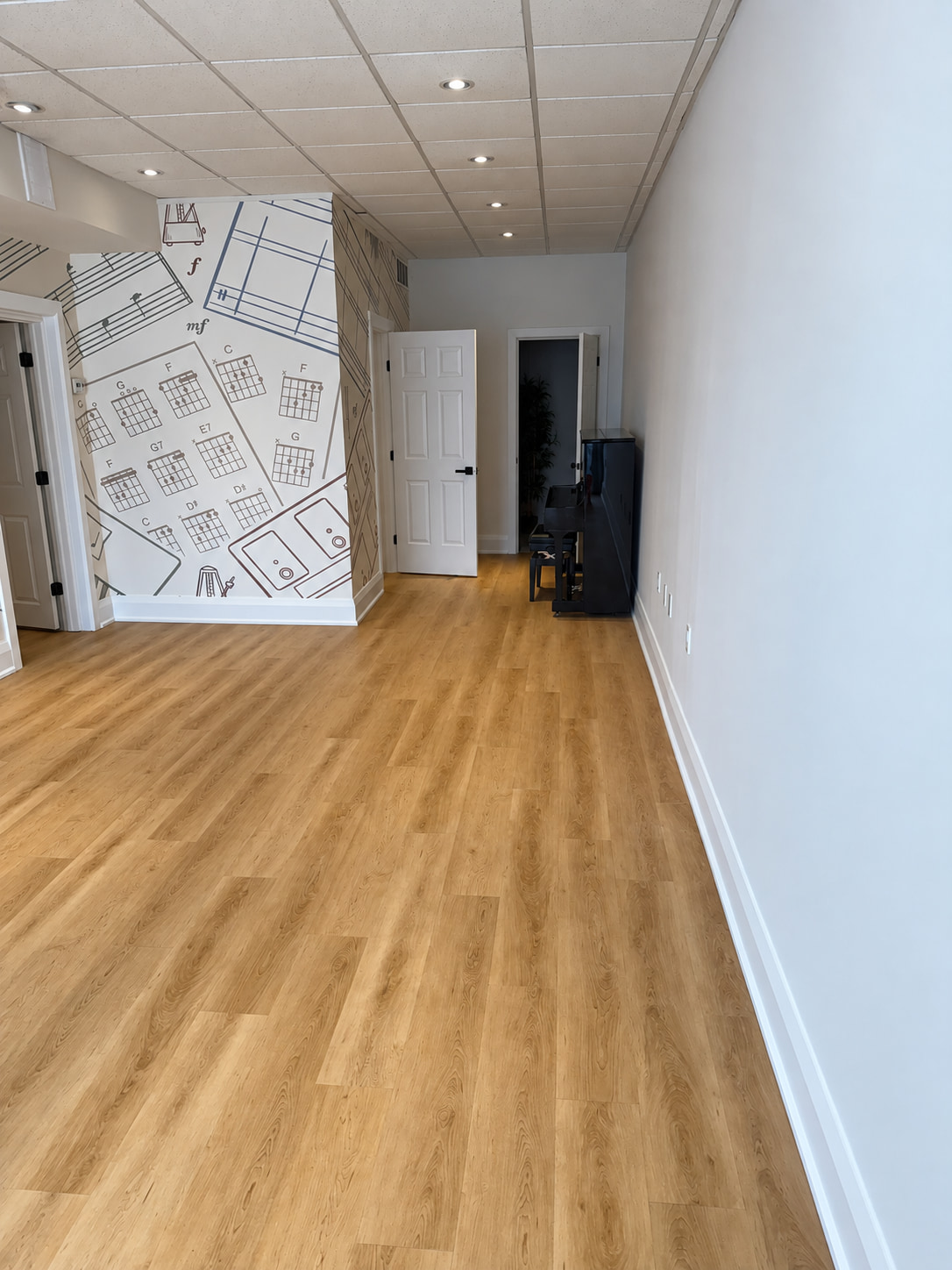 Long music studio room with wood look plank flooring, white walls and an upright piano