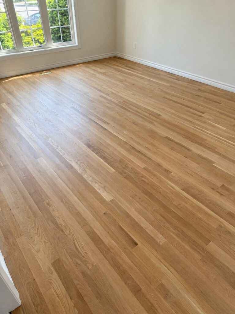 Pale oak strip floor in an empty, bright room with a large window and white baseboards