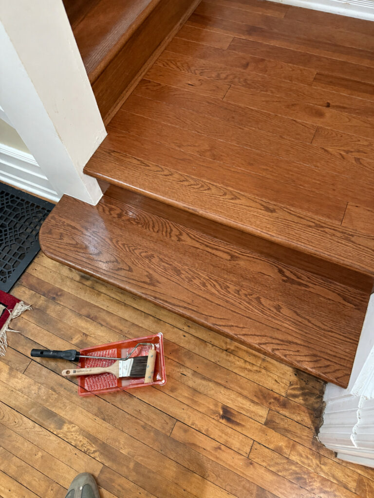 Stained oak stair landing and bottom step above an older wood floor, with painting supplies during the job