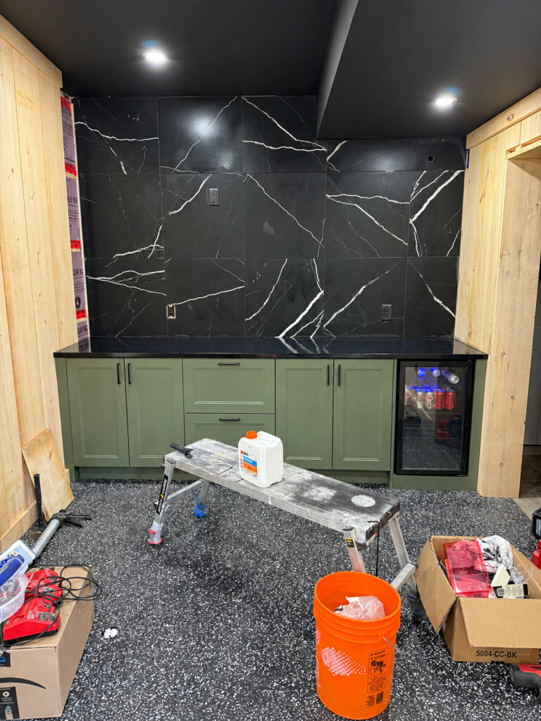 Garage conversion in progress, with a black marble look tile wall, green bar cabinets and a flake floor
