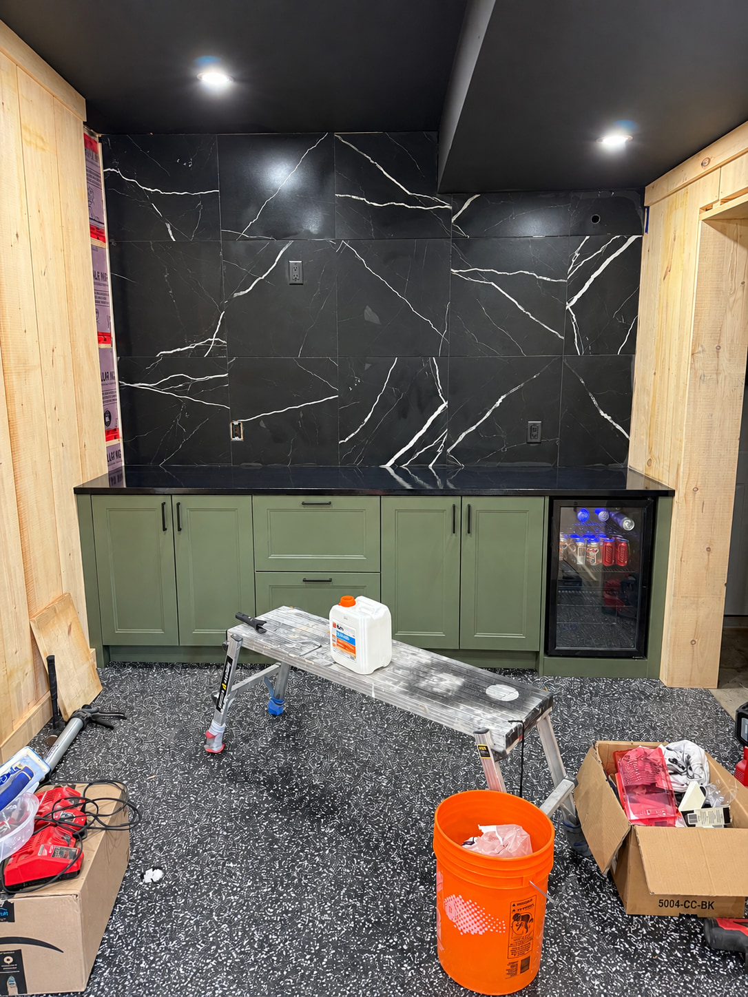 Garage conversion in progress, with a black marble look tile wall, green bar cabinets and a flake floor