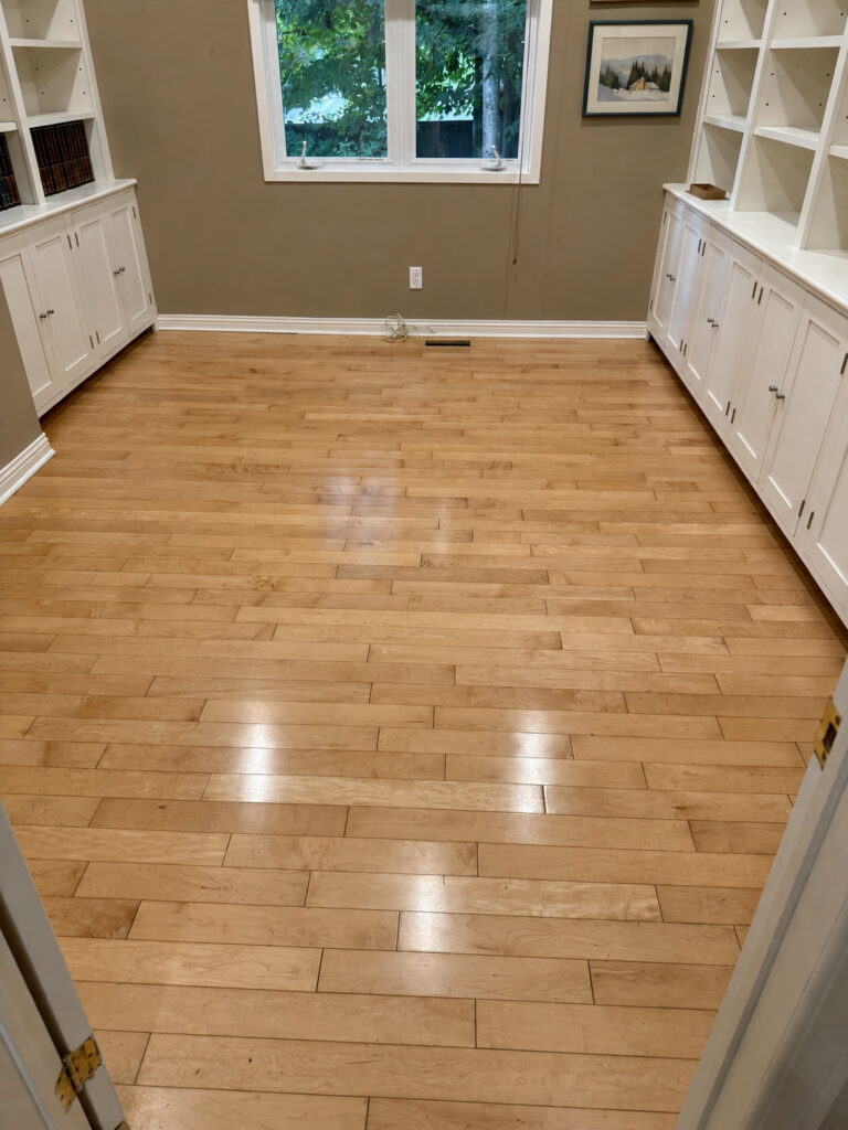 Glossy strip hardwood floor in a den between white built-in cabinets and shelves below a window