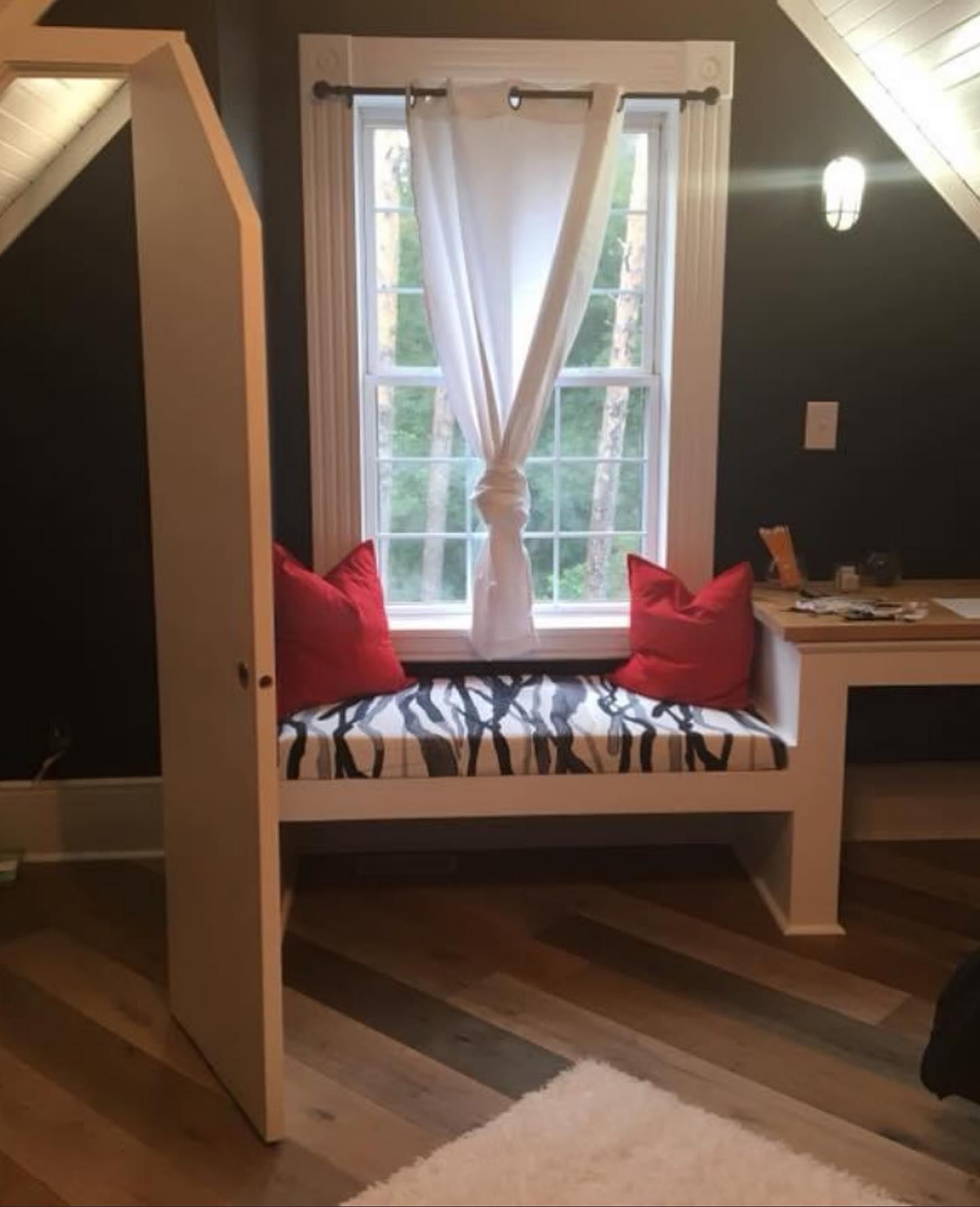 Built-in window seat with red cushions beside a desk, seen through a bedroom door