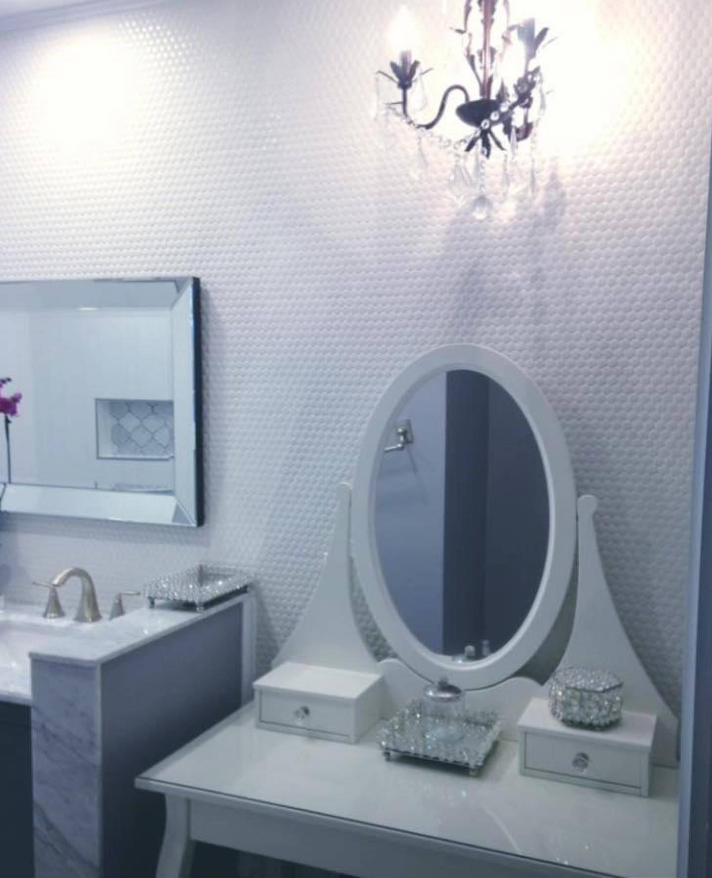 White makeup table with an oval mirror beside a marble-topped vanity and a crystal wall sconce